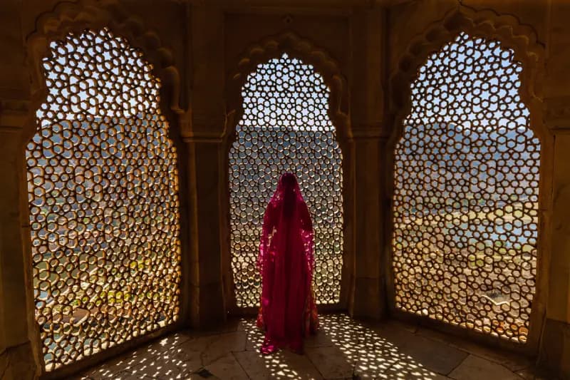 Rajasthan fort, India woman-in-a-red-veiled-sari-standing-before-an-intricately-latticed-arched-window-inside-a-historic-fort-in-rajasthan-india