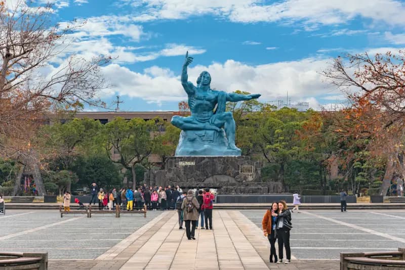 Estátua da Paz de Nagasaki turistas-caminhando-tirando-fotos-em-frente-a-estatua-da-paz-de-nagasaki-no-parque-da-paz-cercada-por-arvores-e-um-predio-comprido-sob-um-ceu-azul-com-nuvens