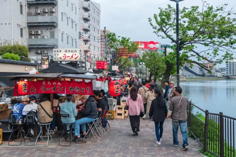 Fukuoka, Japão Barracas de comida de rua (yatai) à beira do rio com pessoas caminhando e jantando, servindo ramen e yakitori, em Fukuoka, Japão