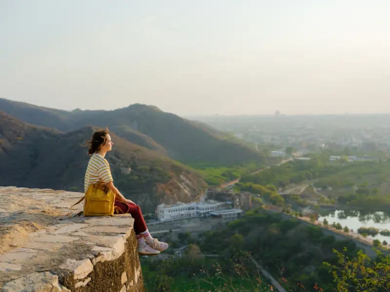 Panoramic view of Jaipur woman-with-a-yellow-backpack-sitting-on-the-edge-of-a-stone-wall-overlooking-a-panoramic-view-of-jaipur-india